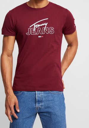 Dunkelrotes T-Shirt mit kurzen Ärmeln mit weißem "Tommy Jeans" Schriftzug und kleinem Logo, kombiniert mit hellblauen Jeans.