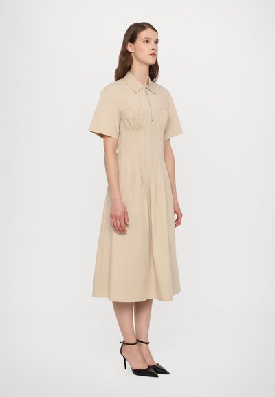 Beige kauluspaita-mekko, jossa on lyhyet hihat, edessä vetoketju ja pleatattu muotoilu. Yhdistetty mustiin kärkitossuihin, joissa on nilkkahihnat.