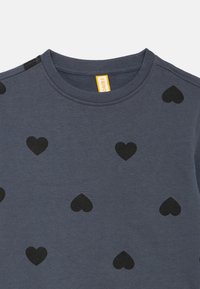 Tamsiai mėlyna medvilninė palaidinė su apvalia iškirpte ir visame paviršiuje juodų širdelių raštu. Ant etiketės geltonais rašmenimis užrašyta „FRIBO“.