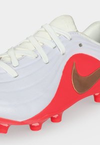 Λευκό και κόκκινο ποδοσφαιρικό παπούτσι Nike με λευκά κορδόνια και μπρονζέ λογότυπο Nike Swoosh στο πλάι.