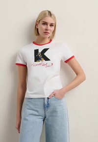 T-shirt branca de mangas curtas com detalhes em vermelho, apresentando uma grande letra 'K' preta e uma personagem de desenho animado com pés amarelos embaixo.