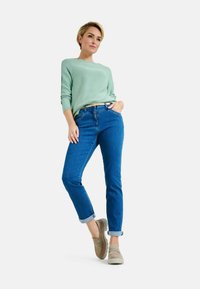 Pullover verde chiaro, jeans blu e scarpe slip-on beige. I jeans hanno gambe affusolate, orli arrotolati e un design classico a cinque tasche.