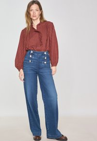 Blusa a scacchi rossa e marrone con dettagli a balze, abbinata a pantaloni in denim blu a vita alta con grandi bottoni e design a gamba larga.