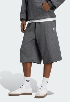 Persona con pantalones cortos Adidas gris oscuro, sudadera con capucha a juego, calcetines blancos Adidas y zapatillas blancas, de pie frente a un fondo claro.