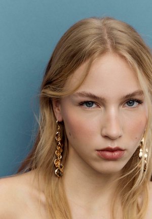 Junge Frau mit blondem Haar und blauen Augen, die lange goldene Kettenohrringe trägt und vor einem einfarbigen blauen Hintergrund posiert.