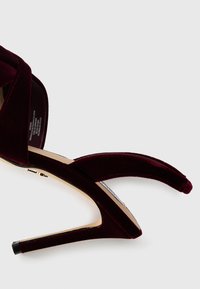 Bordo plišani sandali s visokom petom, otvorenim prstima i preklapajućim trakama. Imaju elegantnu stiletto petu i glatku, sjajnu potplat.