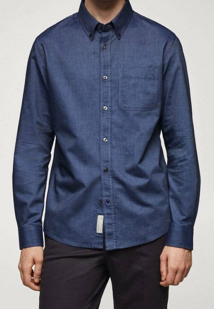 Homme portant une chemise à manches longues bleu foncé boutonnée avec une poche poitrine et un pantalon foné, debout les mains détendues le long du corps.