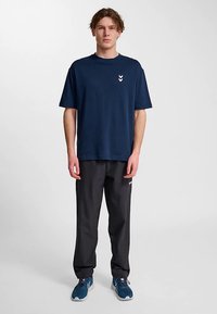 Mörkblå bomulls-t-shirt med korta ärmar och en vit chevronlogotyp. Kombinerad med svarta byxor och blåa sportskor.