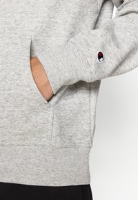 Grauer Fleece-Sweatshirt mit strukturierter Oberfläche, Fronttasche, gerippten Bündchen und einem Logo-Patch am Ärmel.