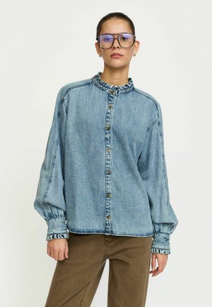 Koszula z denimu w jasnoniebieskim kolorze z falbankowym kołnierzem, zapinana na guziki z przodu i z bufiastymi rękawami. Posiada kontrastowe przeszycia oraz elastyczne mankiety.