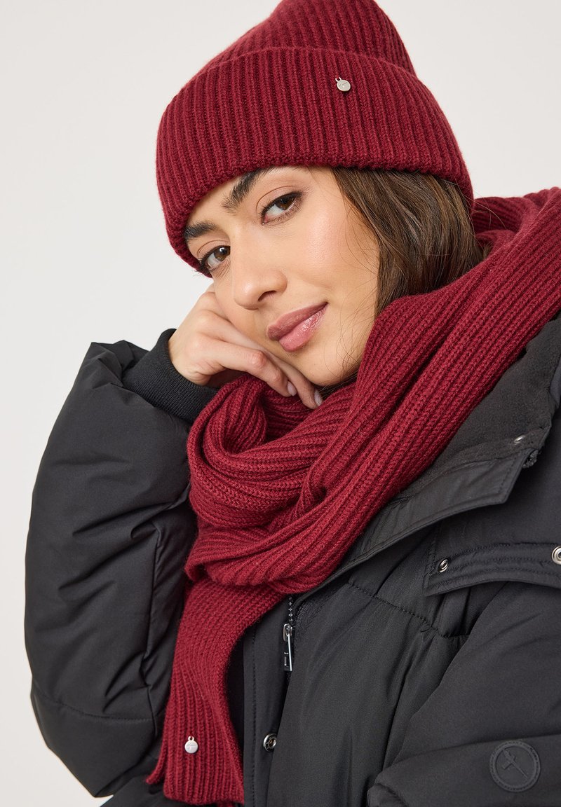 Cappello a coste rosso e sciarpa coordinata drappeggiata attorno al collo, abbinati a un giubbotto imbottito nero con dettagli e cerniere argentate.