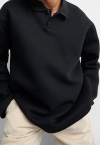 Svart långärmad polo med krage och knappslå, tillverkad av ett glatt tyg, i kombination med krämfärgade byxor.