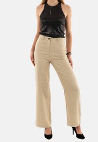 Pantaloni beige a gamba larga con finitura testurizzata, tasche frontali e un bottone dorato. Abbinati a un top halter nero e tacchi neri.