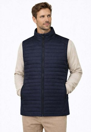 Uomo che indossa un gilet trapuntato blu navy con zip sopra una camicia beige a maniche lunghe, con le mani nelle tasche del gilet, che guarda leggermente di lato.
