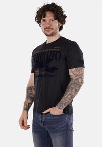 Camiseta de algodón negra de manga corta con un diseño gráfico que presenta la palabra "Tornado" y motivos militares en azul oscuro y naranja.