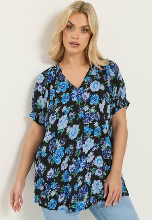 Blondynka w czarnej bluzce z krótkim rękawem z niebiesko-białym kwiatowym wzorem, w jasnoniebieskich dżinsach, stojąca na tle jednolitej ściany.