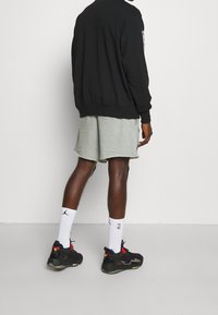 Sudadera negra, pantalones cortos gris claro, medias de baloncesto blancas y zapatillas deportivas negras con acentos rojos. Vista trasera, de pie sobre una superficie lisa.