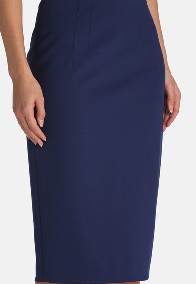 Woman wearing a fitted knee-length navy blue skirt with subtle seam details, hands resting by her sides against a plain background.
