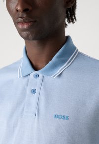 Svetlomodrá polo tričko z textúrovaného materiálu, s rebrovým golierom s bielymi pruhmi a malým modrým logom "BOSS" na hrudi.