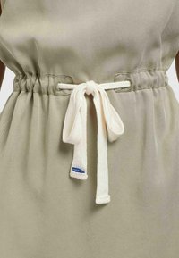 Robe en tissu beige avec une ceinture en corde blanche nouée en nœud papillon à la taille, ornée d'une petite étiquette en tissu bleu à l'extrémité de la ceinture.