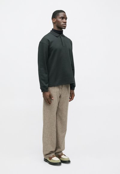 Άνδρας που στέκεται φορώντας μαύρο μακρύ μανίκι πόλο, μπεζ εκρού σκούρα παντελόνια με φαρδιά πόδια και μπεζ και καφέ μοκέτες με μαύρες σόλες.