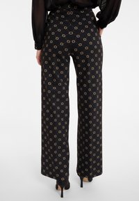 Wide-leg black pants with a gold geometric pattern, featuring a high waist and smooth fabric. Styled with pointed black heels.