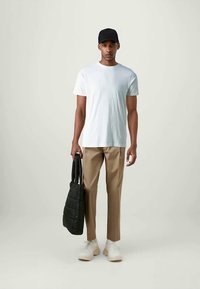 Άνδρας που φοράει μαύρο καπέλο, άσπρη μπλούζα, μπεζ παντελόνι και άσπρα sneakers, κρατώντας μια μαύρη τσάντα με επένδυση σε έναν απλό φόντο.