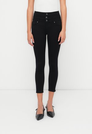 Femme portant un jean skinny noir taille haute avec trois boutons et chevilles raccourcies, associé à des talons slingback noirs pointus.