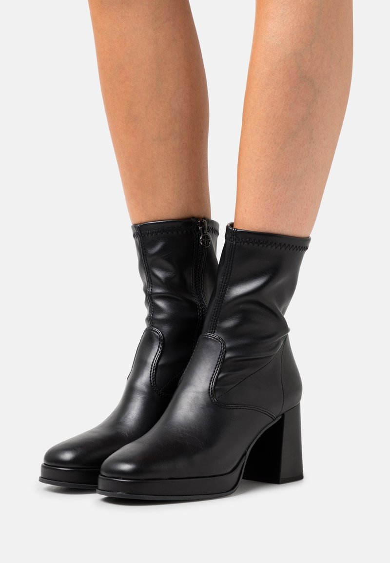 Bottes en cuir noir mi-mollet avec talons carrés et fermetures éclair latérales portées sur jambes nues sur fond blanc.