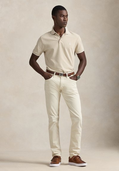 Μπεζ ριγέ πόλο πουκάμισο, κρεμ τζιν και καφέ αθλητικά παπούτσια. Το μοντέλο στέκεται με τα χέρια στις τσέπες, αναδεικνύοντας ένα καθημερινό ντύσιμο.