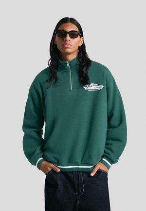 Green fleece quarter-zip sweatshirt with white accents, relaxed fit, and a logo on the chest. Paired with dark denim pants.