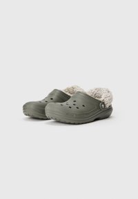 Ein Paar olivgrüne Clogs mit offenen Löchern oben und beigem, flauschigem Innenfutter, vor einem weißen Hintergrund.