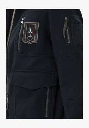 Particolare del maniche e del petto di una giacca nera con molte tasche con zip e una patch che presenta un arco stilizzato e tre strisce colorate.