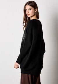 Czarny, oversize sweter z długimi rękawami, z geometrycznym wzorem w srebrnych zdobieniach z przodu. Dzianinowa tekstura; gładkie wykończenie.