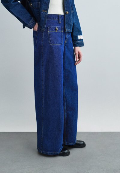 Pantalon en denim bicolore à jambes larges avec poches à rabat à l'avant, porté avec une veste en denim bleu foncé et des chaussures noires sur un sol gris.