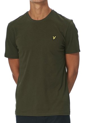 Hombre con camiseta de manga corta verde oliva con un pequeño logo amarillo de un pájaro en el pecho izquierdo, de pie con las manos detrás de la espalda.