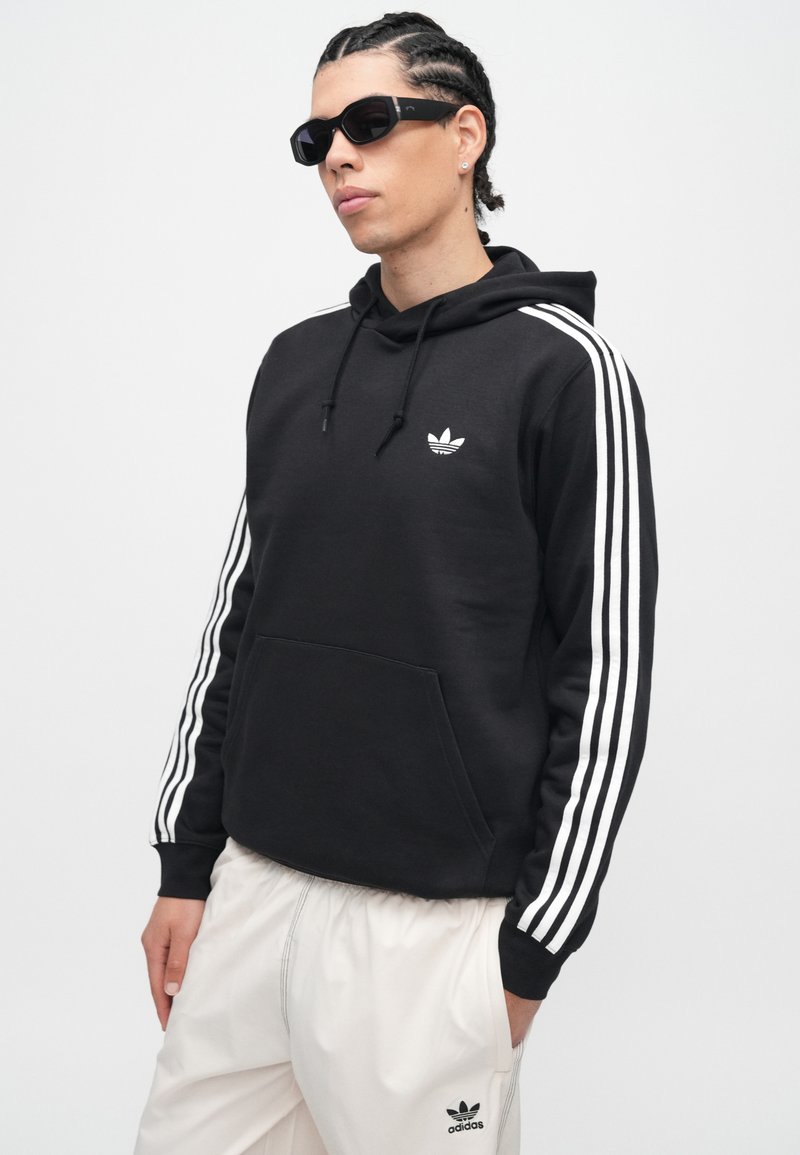 Giovane con capelli intrecciati che indossa una felpa nera Adidas con strisce bianche e pantaloni bianchi Adidas, posa con le mani nelle tasche.