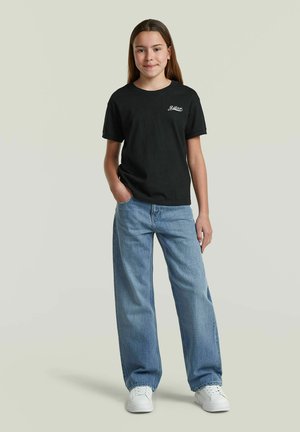 Jeune personne debout, main dans la poche, portant un t-shirt noir, un jean bleu ample et des baskets blanches sur fond uni.
