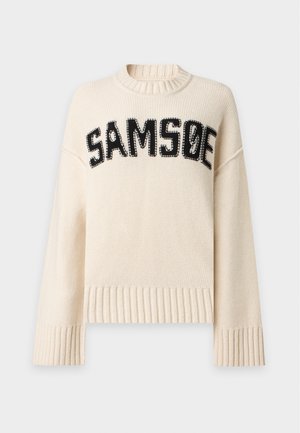 Mujer con cabello largo y negro lleva un suéter crema de gran tamaño con el texto "SAMSO", pantalones caqui sueltos y zapatos marrón oscuro, de pie con una mano en el bolsillo.