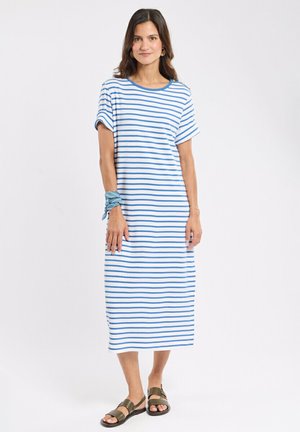Femme portant une robe midi blanche à rayures horizontales bleues, des sandales vertes et une écharpe bleue nouée autour de son poignet, debout sur un fond blanc.