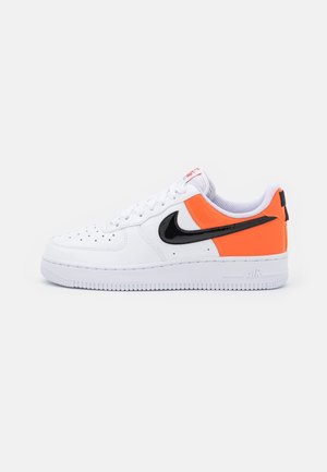 Baskets basses blanches avec un swoosh noir de Nike, un talon et un panneau latéral orange vif, des lacets blancs et une boîte à orteils perforée.