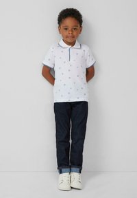 Chemise polo blanche avec des motifs floraux bleus et un col, associée à un jean en denim sombre et des baskets blanches. L'enfant se tient avec les mains derrière le dos.