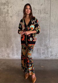 Γυναίκα που στέκεται μπροστά σε τσιμεντένιο τοίχο, φοράει μαύρο κάρντιγκαν με floral σχέδια και ταιριαστό παντελόνι με φαρδιά μπατζάκια, και καφέ μυτερά παπούτσια.