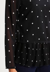 Blusa nera con maniche lunghe, caratterizzata da pois bianchi, una sezione inferiore plissettata e una texture in tessuto leggero e trasparente.