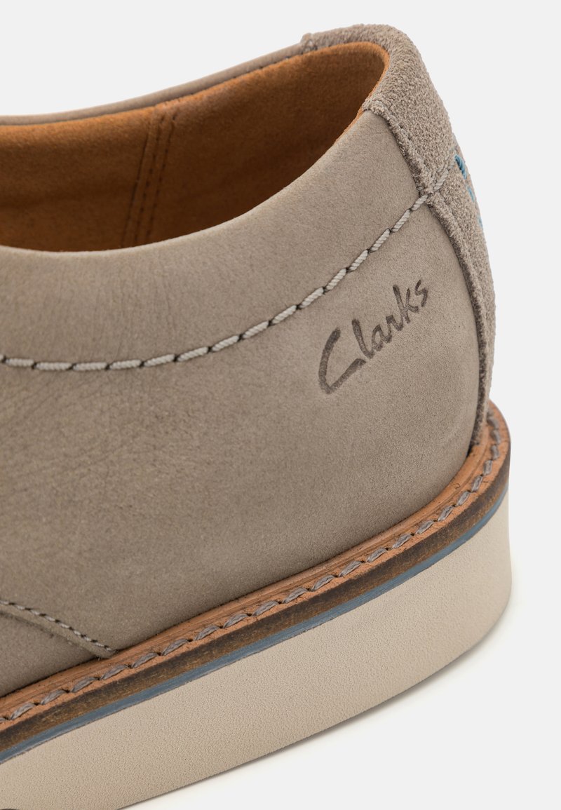 Zapato de ante gris claro con una parte superior lisa, costuras blancas en contraste y una suela de goma beige. Presenta el logo "Clarks" cerca del talón.