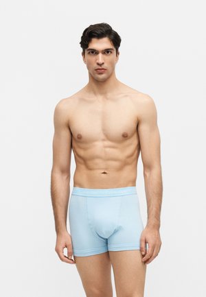 Zadbany młody mężczyzna o ciemnych włosach stoi i nosi jasnoniebieskie bokserki Calvin Klein na jednolitym jasnym tle.