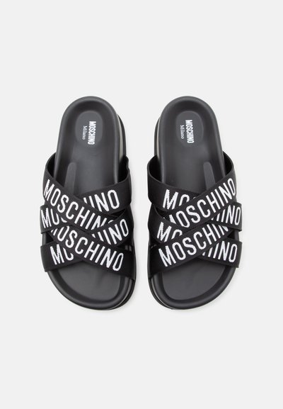 Paire de sandales slides noires Moschino avec des lanières croisées comportant le texte blanc Moschino sur chaque lanière.