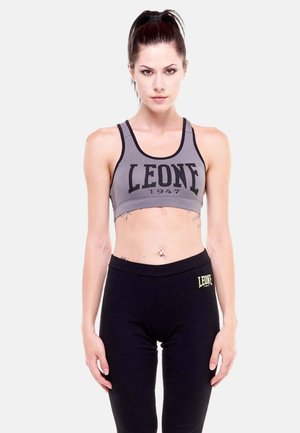 Femme aux cheveux foncés en queue de cheval, portant un soutien-gorge de sport gris et un legging noir, tous deux de la marque "LEONE 1947", debout sur un fond blanc.