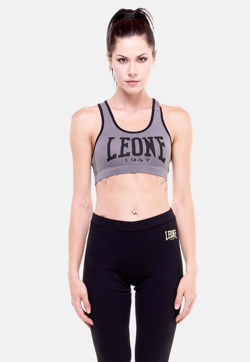 Femme aux cheveux foncés en queue de cheval, portant un soutien-gorge de sport gris et un legging noir, tous deux de la marque "LEONE 1947", debout sur un fond blanc.