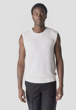 Fiatal férfi ujjatlan fehér texturált kötött felsőben és fekete cargo nadrágban, szürke egyszínű háttér előtt állva.